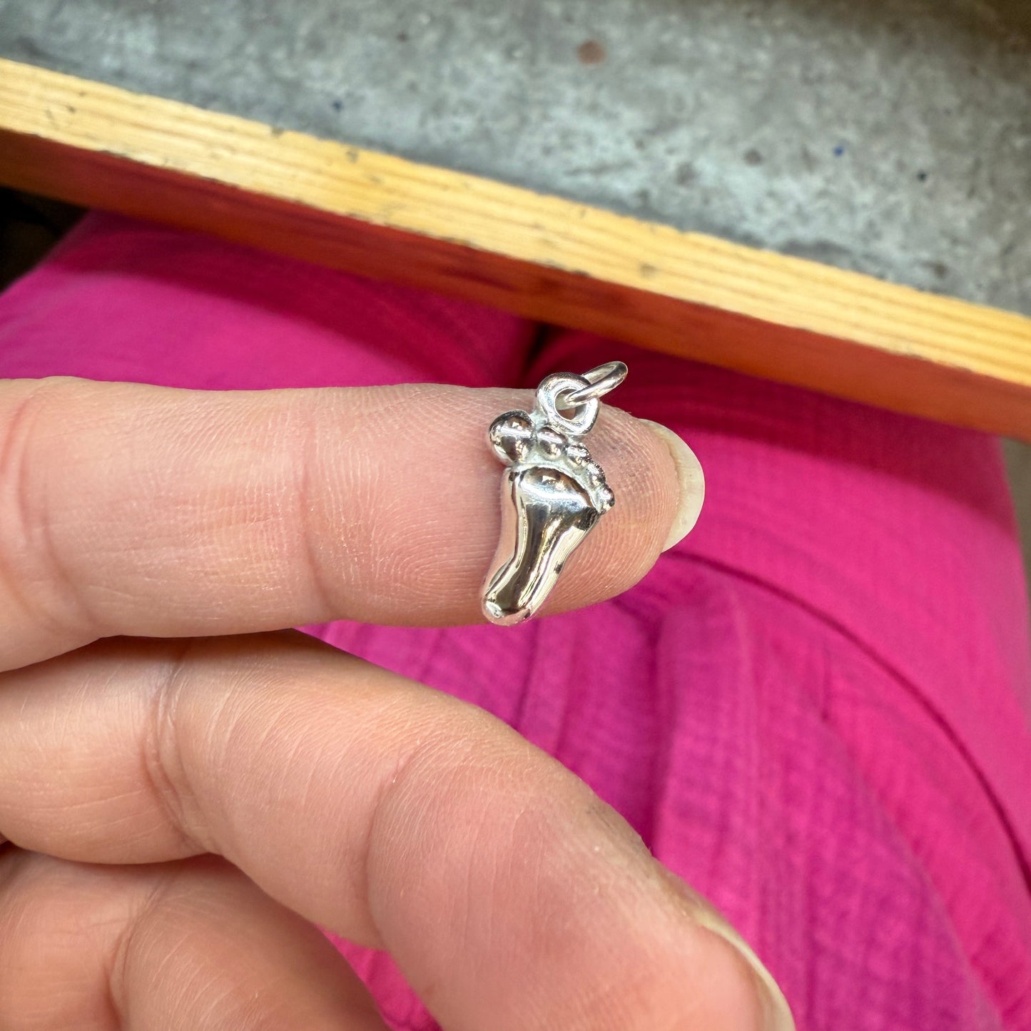 A small silver baby foot charm being shown on a fingertip for size reference.